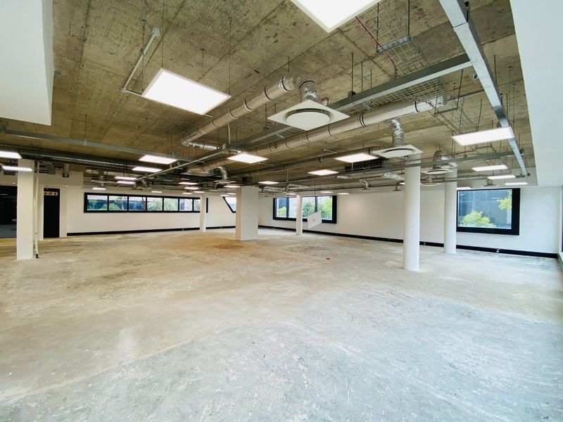 Spacious open-plan commercial office space featuring an industrial aesthetic with exposed concrete ceilings, HVAC ducting, and large windows providing ample natural light.