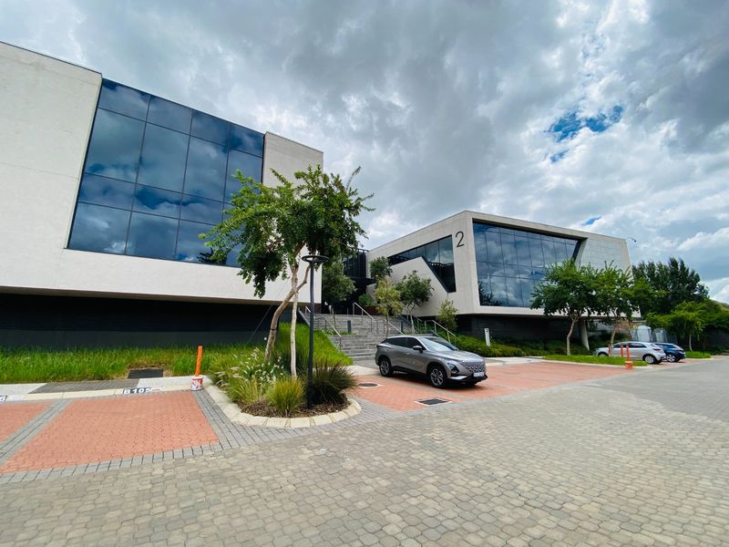 Modern commercial office building featuring a glass facade, paved parking area, and landscaped grounds with concrete access stairs.