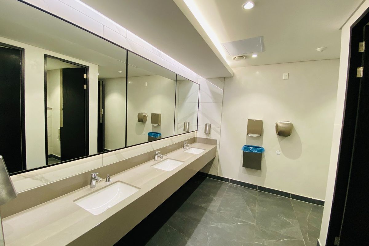 Modern commercial bathroom featuring a triple vanity with stone-look countertop, large integrated mirrors, recessed lighting, and wall-mounted fixtures.
