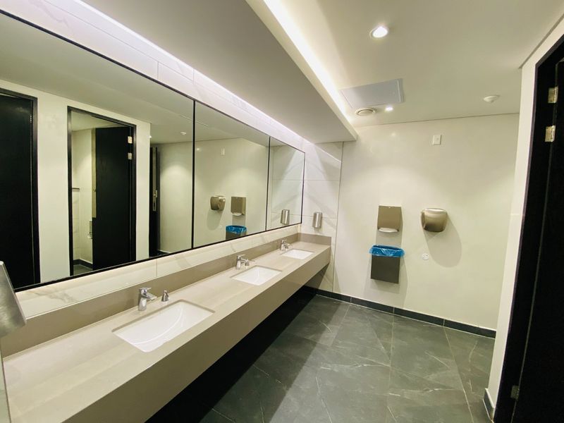 Modern commercial bathroom featuring a triple vanity with stone-look countertop, large integrated mirrors, recessed lighting, and wall-mounted fixtures.