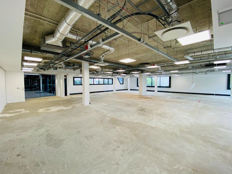 Spacious open-plan commercial office area featuring an industrial aesthetic with exposed concrete ceilings, ducting, and large windows providing ample natural light.