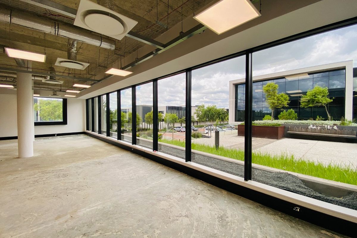 A modern, open-plan office space featuring an industrial aesthetic with exposed ceilings, concrete floors, and expansive floor-to-ceiling windows overlooking a landscaped courtyard.