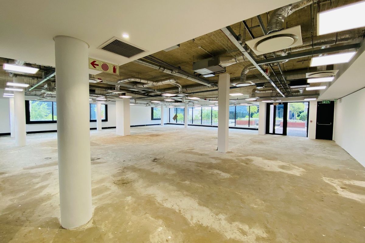 A large, open-plan commercial office space featuring an industrial aesthetic with exposed ceilings, ducting, and large windows providing ample natural light.