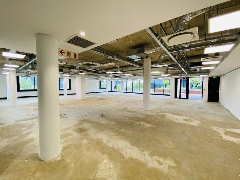 A large, open-plan commercial office space featuring an industrial aesthetic with exposed ceilings, ducting, and large windows providing ample natural light.