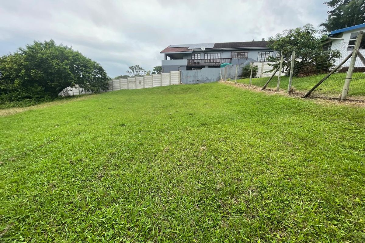 A view of a vacant residential plot featuring a grassy slope, perimeter fencing, and a neighboring house in the background.