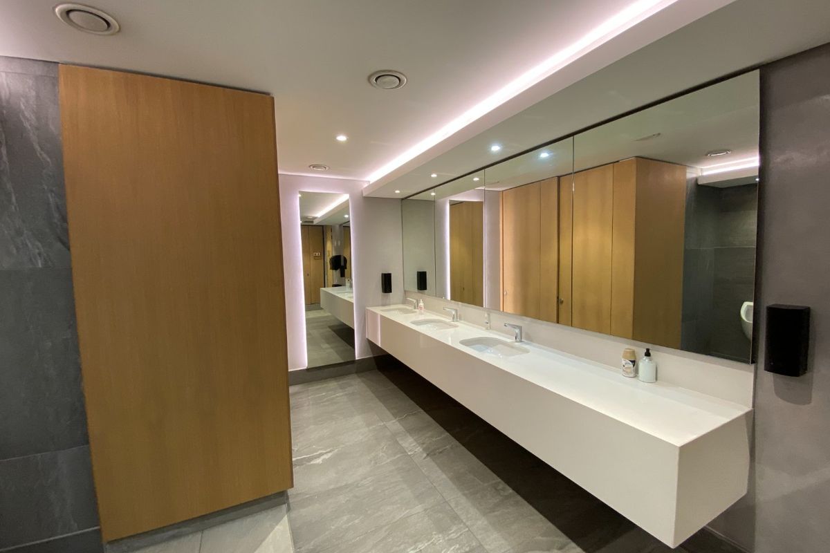Modern commercial bathroom featuring a sleek double vanity with integrated sinks, large wall-to-wall mirror, and sophisticated recessed and cove lighting.