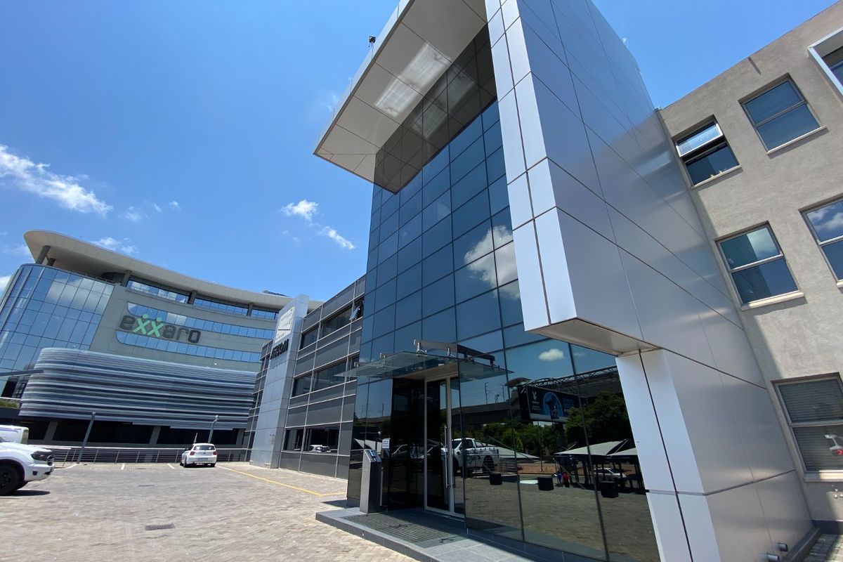 Modern commercial office building exterior featuring a glass curtain wall facade, metal cladding, and a paved parking area.