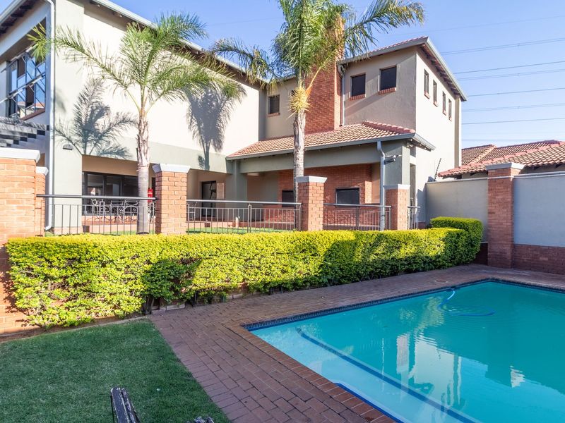 A well-maintained outdoor pool area featuring brick paving, a perimeter wall with metal railings, and a covered patio space.
