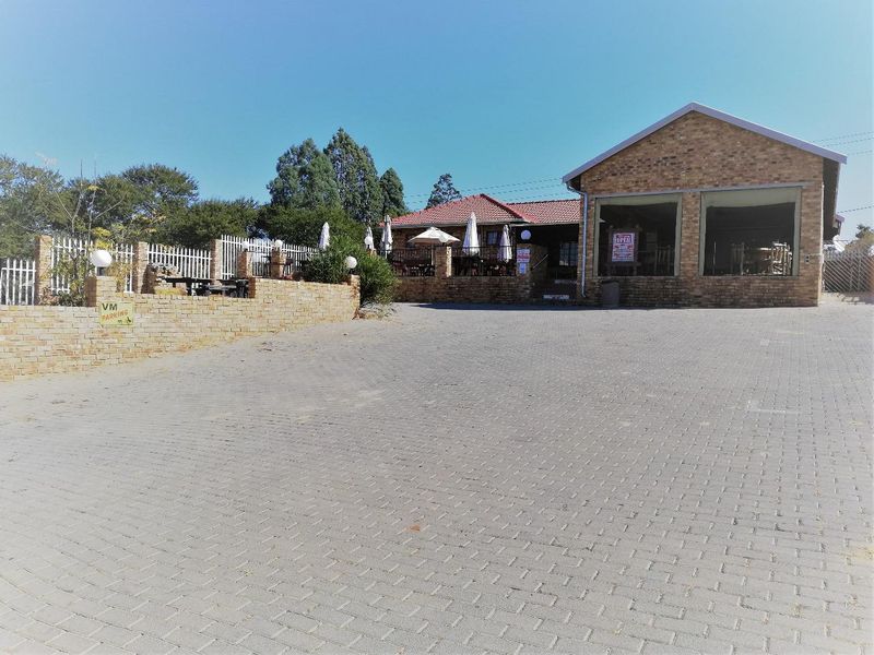 Front exterior view of a brick building featuring a large paved driveway, boundary wall, and a covered patio area.