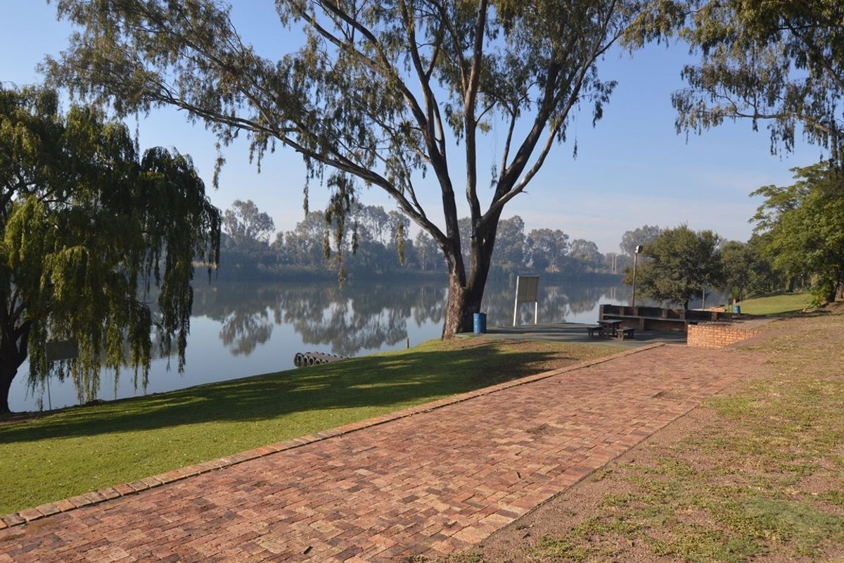 A scenic riverside garden area featuring brick paved pathways, mature trees, and a built-in brick braai facility overlooking the water.