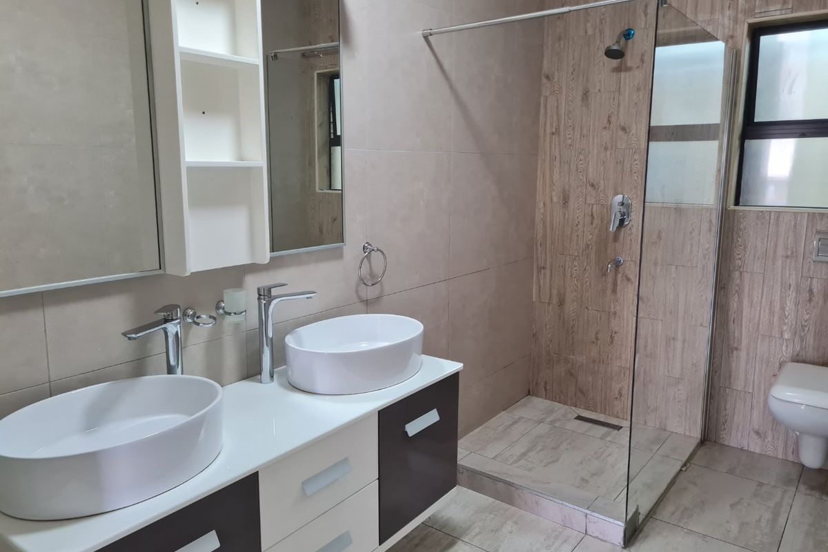Modern bathroom featuring a double vanity with vessel sinks, a spacious walk-in shower with glass screen, and a wall-mounted toilet with neutral tiling.