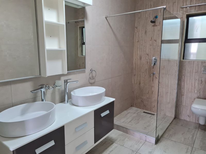 Modern bathroom featuring a double vanity with vessel sinks, a spacious walk-in shower with glass screen, and a wall-mounted toilet with neutral tiling.