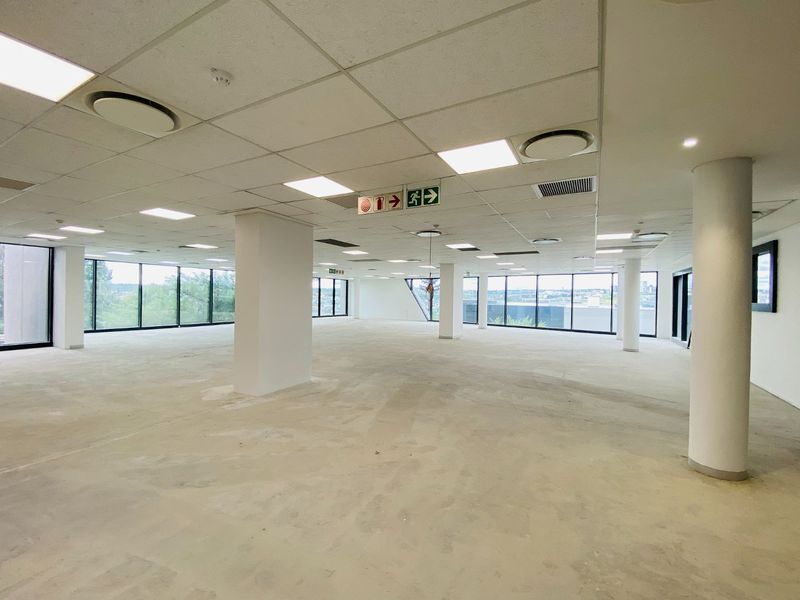 A spacious, open-plan commercial office area featuring large windows, suspended ceilings with recessed lighting, and structural columns, currently in a shell condition.