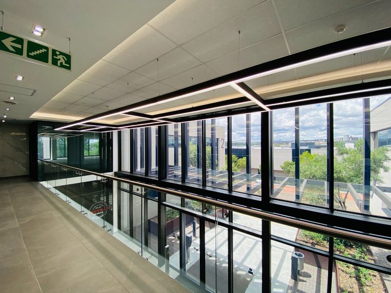 Modern commercial hallway featuring a glass balustrade, large format floor tiles, and expansive floor-to-ceiling windows overlooking the exterior courtyard.