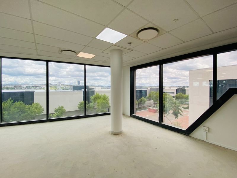 An empty, open-plan office space featuring large floor-to-ceiling windows, a suspended ceiling with recessed lighting, and a central structural column.