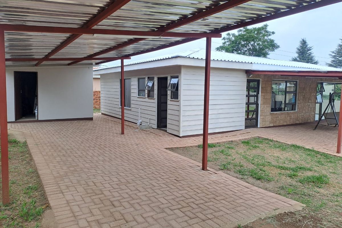 Rear exterior view featuring a paved driveway, covered carport area, and a house with stone cladding and corrugated metal roofing.