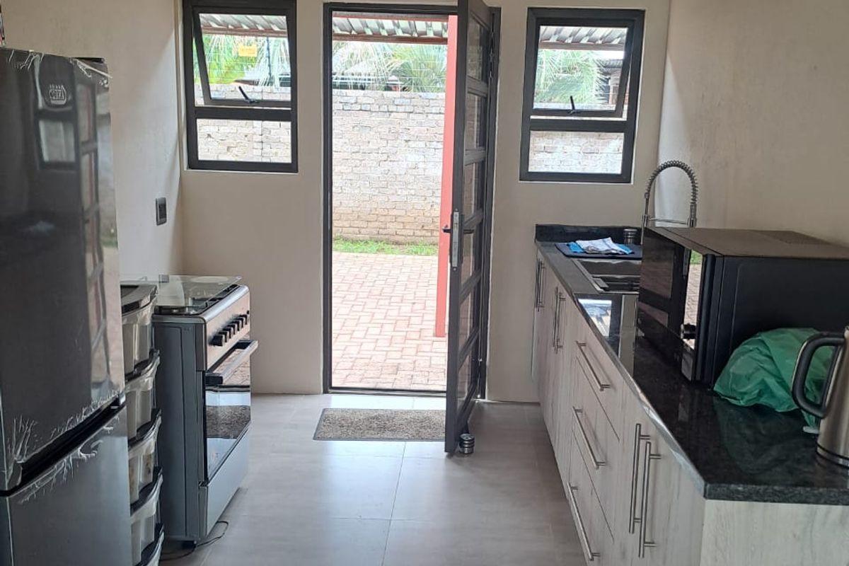 A modern galley-style kitchen featuring granite countertops, light wood-finish cabinetry, tiled flooring, and direct access to the rear outdoor area.