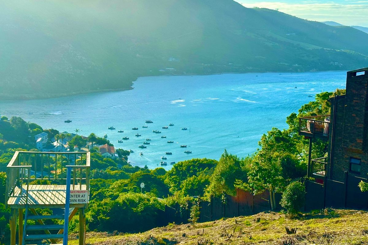 A scenic elevated view overlooking a coastal bay with boats, featuring a wooden viewing platform on the foreground slope.
