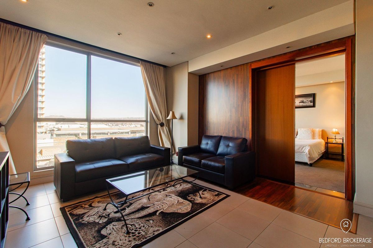 A modern living room featuring large floor-to-ceiling windows, recessed lighting, and a wood-paneled feature wall, with an adjacent bedroom accessible via a sliding door.