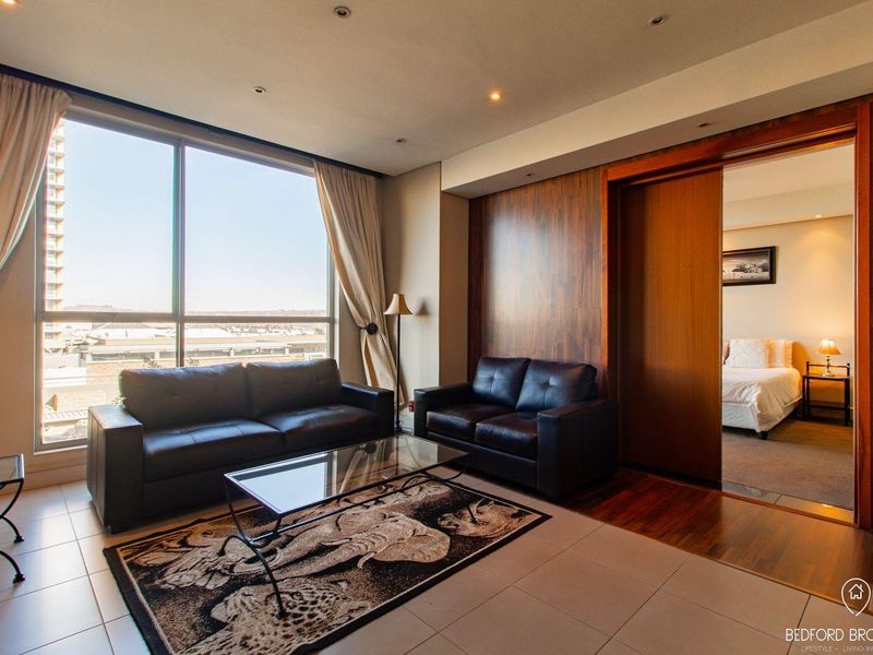 A modern living room featuring large floor-to-ceiling windows, recessed lighting, and a wood-paneled feature wall, with an adjacent bedroom accessible via a sliding door.
