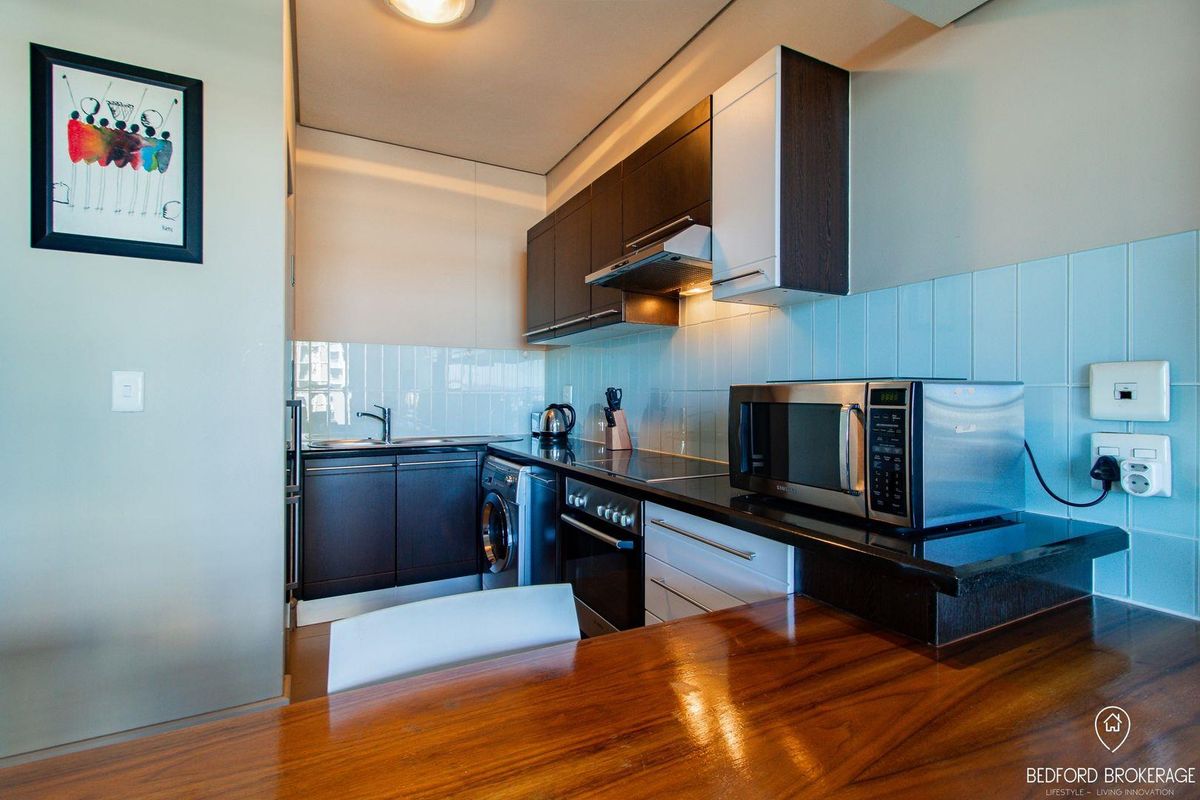 Compact modern kitchen featuring dark cabinetry, stone countertops, integrated appliances, and a light blue tiled splashback.