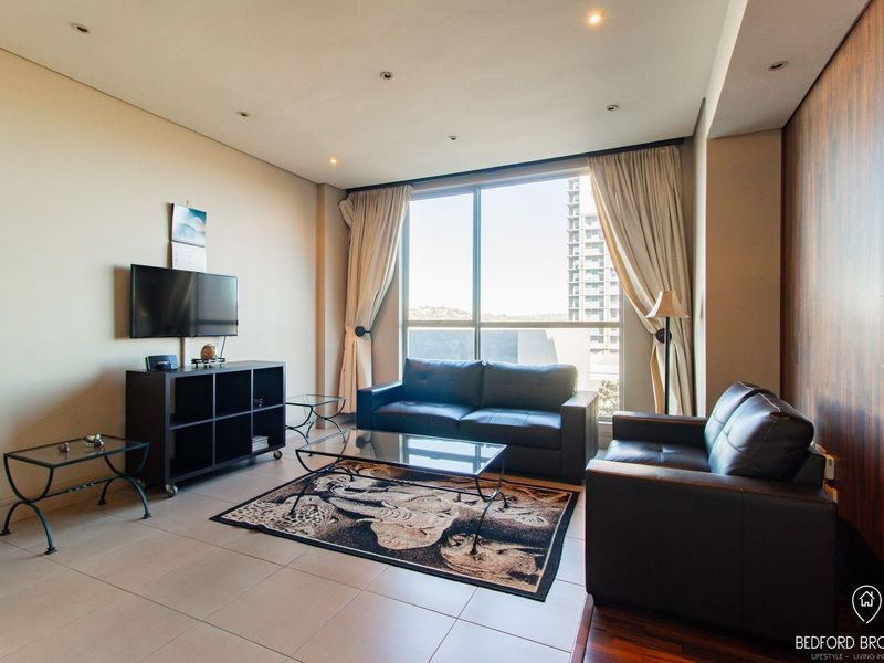 A well-lit living room featuring large floor-to-ceiling windows, tiled flooring, and a modern wood-panelled feature wall.