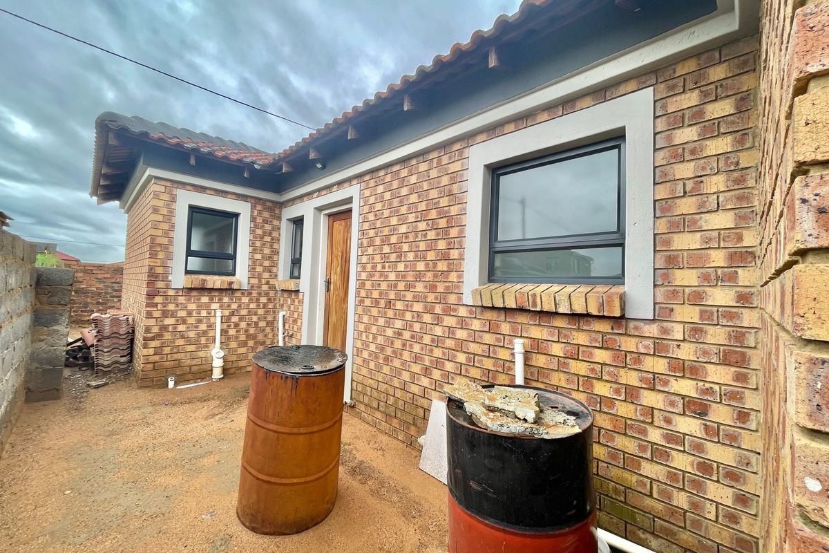 Rear exterior view of a face brick house featuring tiled roofing, aluminium window frames, and unfinished ground area.