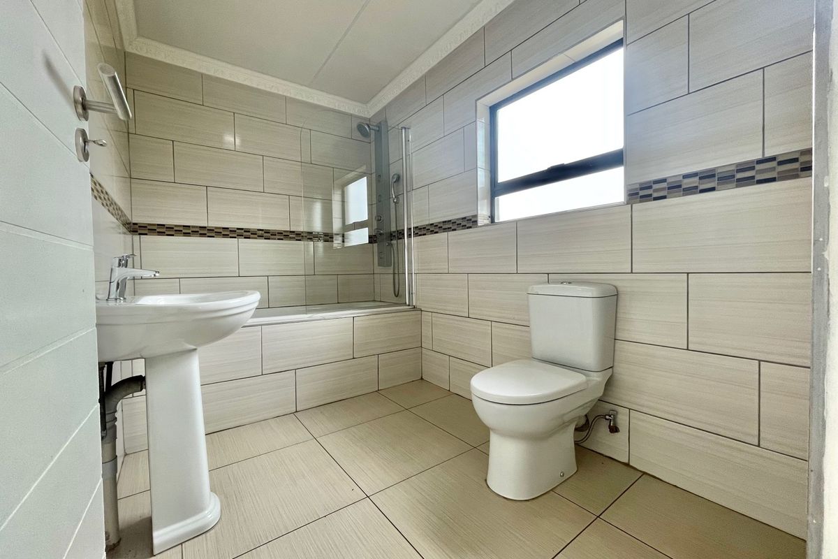 A clean, modern bathroom featuring a pedestal basin, toilet, and a bathtub with an integrated shower and glass screen, finished with neutral wall tiles.