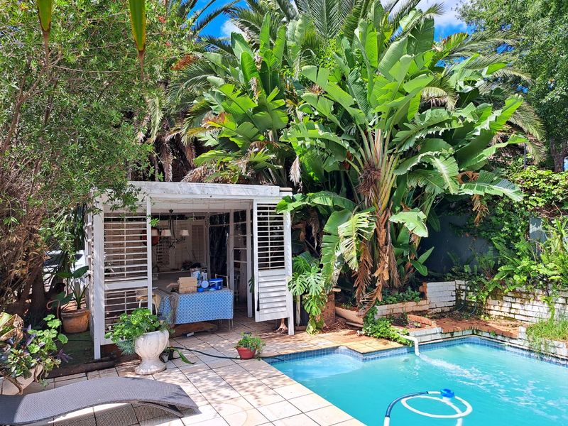 Outdoor pool area featuring a tiled deck, a covered patio structure, and established garden landscaping with a brick retaining wall.