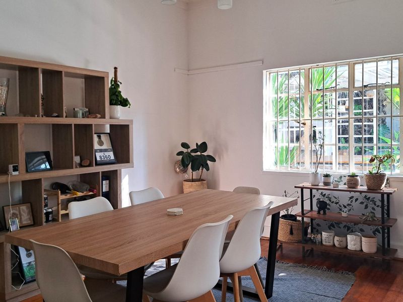 A bright dining room featuring warm hardwood flooring and a large window fitted with security bars, providing natural light and a view of the garden.