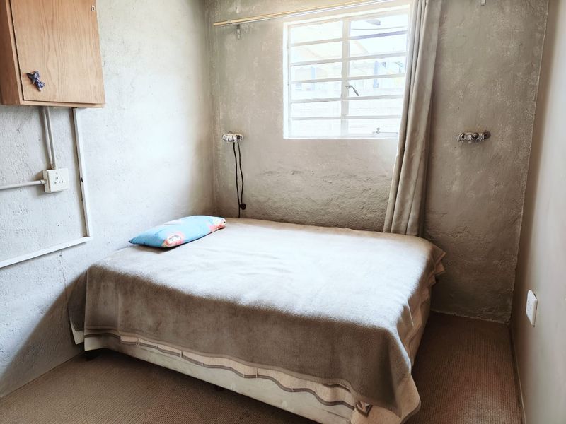 A compact bedroom featuring rough-textured walls, a window with security bars, and carpeted flooring, requiring cosmetic updates to improve the overall finish.