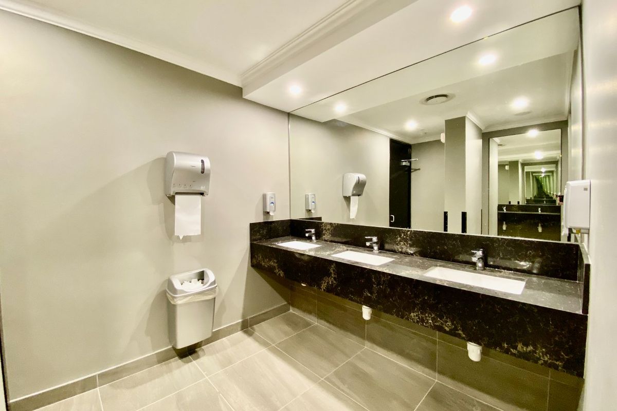A modern commercial bathroom featuring a triple vanity with a stone countertop, large wall mirror, recessed lighting, and neutral tiled flooring.