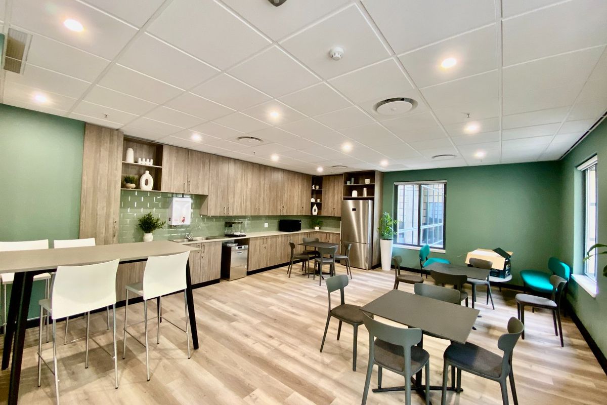 A modern commercial kitchen and breakroom area featuring wood-finish cabinetry, integrated appliances, subway tile backsplash, and a suspended ceiling with recessed lighting.