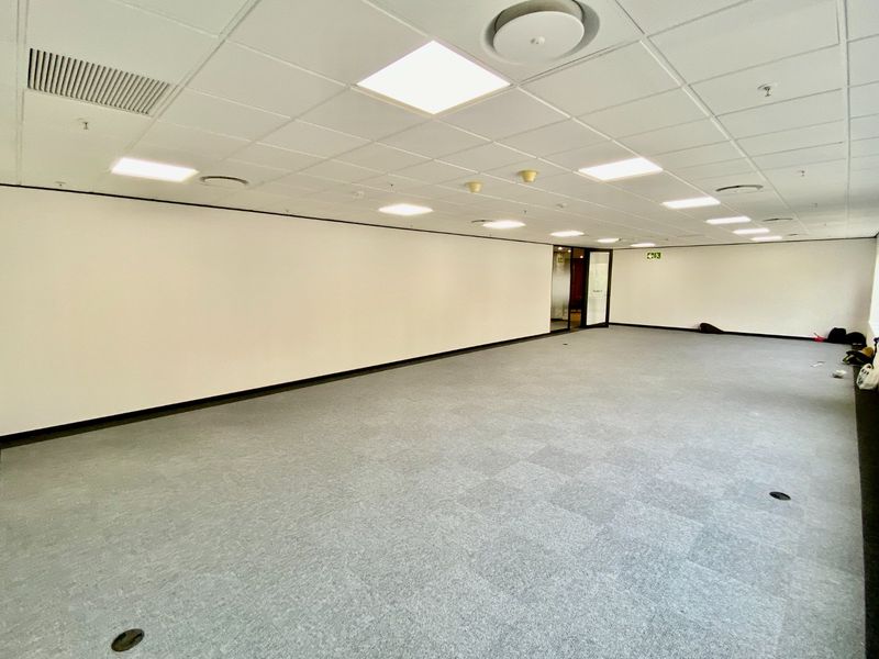A spacious, empty commercial office area featuring a suspended ceiling with recessed lighting, carpeted flooring, and integrated air conditioning vents.