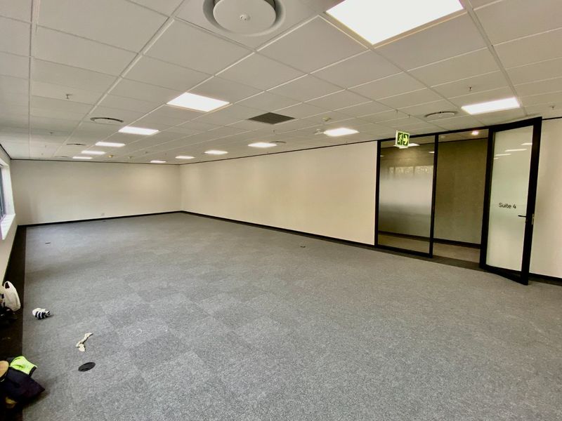 A spacious, empty commercial office suite featuring neutral carpet tiles, a suspended ceiling with recessed lighting, and a modern glass partition entrance.