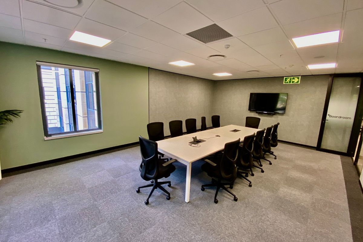A professional boardroom featuring a large meeting table, carpeted flooring, suspended ceiling with recessed lighting, and a wall-mounted television.