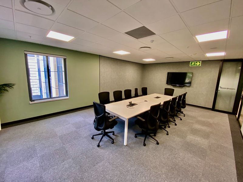 A professional boardroom featuring a large meeting table, carpeted flooring, suspended ceiling with recessed lighting, and a wall-mounted television.