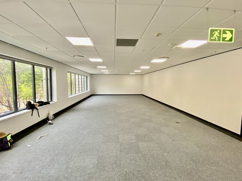 A spacious, empty commercial office area featuring neutral carpet tiles, a suspended ceiling with recessed lighting, and large windows providing natural light.