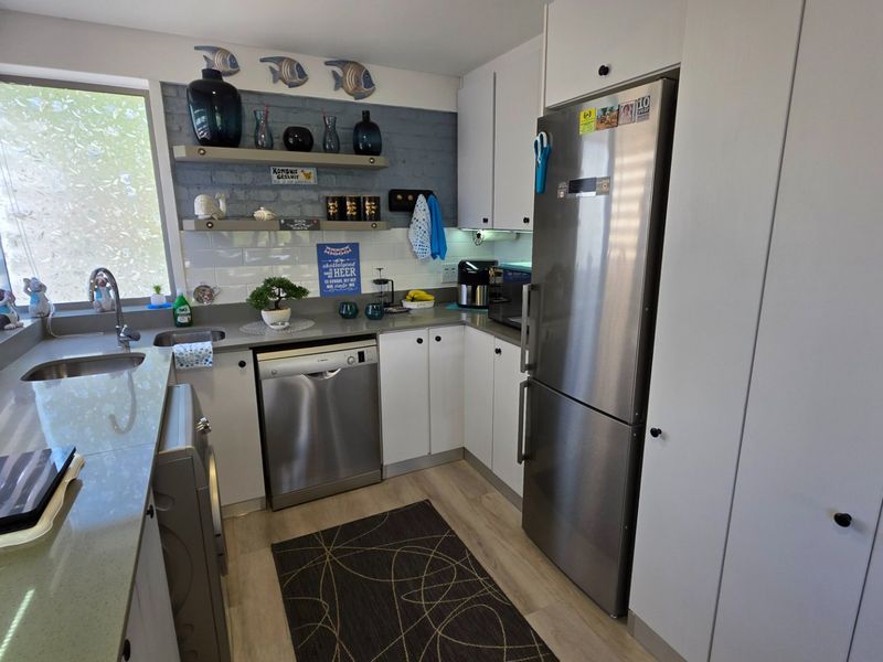 A functional kitchen featuring modern white cabinetry, stone countertops, integrated dishwasher, stainless steel refrigerator, and a stylish tiled backsplash with floating shelving.