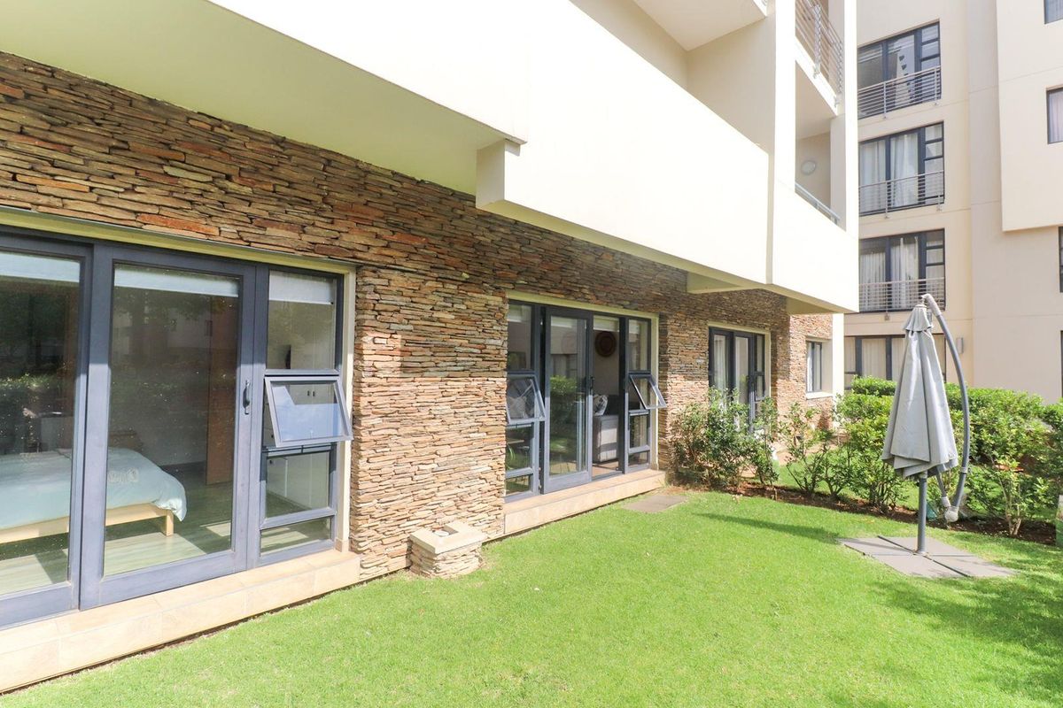 A well-maintained rear exterior view of the apartment featuring stone cladding, modern aluminium sliding doors, and a private garden area.