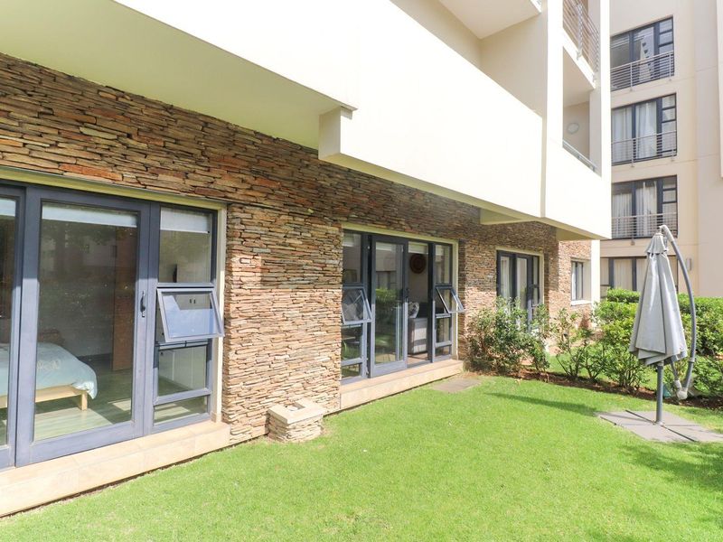 A well-maintained rear exterior view of the apartment featuring stone cladding, modern aluminium sliding doors, and a private garden area.