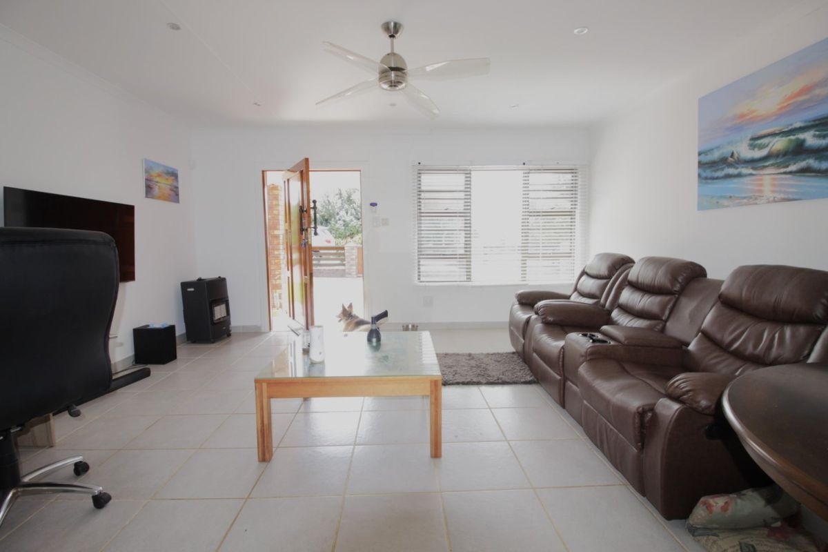 A bright living room featuring tiled flooring, a ceiling fan, and large windows with venetian blinds, providing a functional and well-lit space.