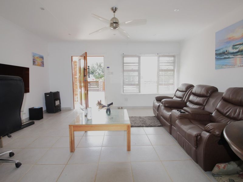 A bright living room featuring tiled flooring, a ceiling fan, and large windows with venetian blinds, providing a functional and well-lit space.