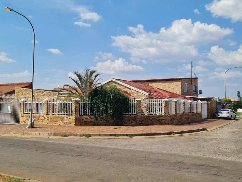 Corner property featuring a face brick facade, tiled roof, and secure perimeter wall with metal fencing, situated on a paved street.