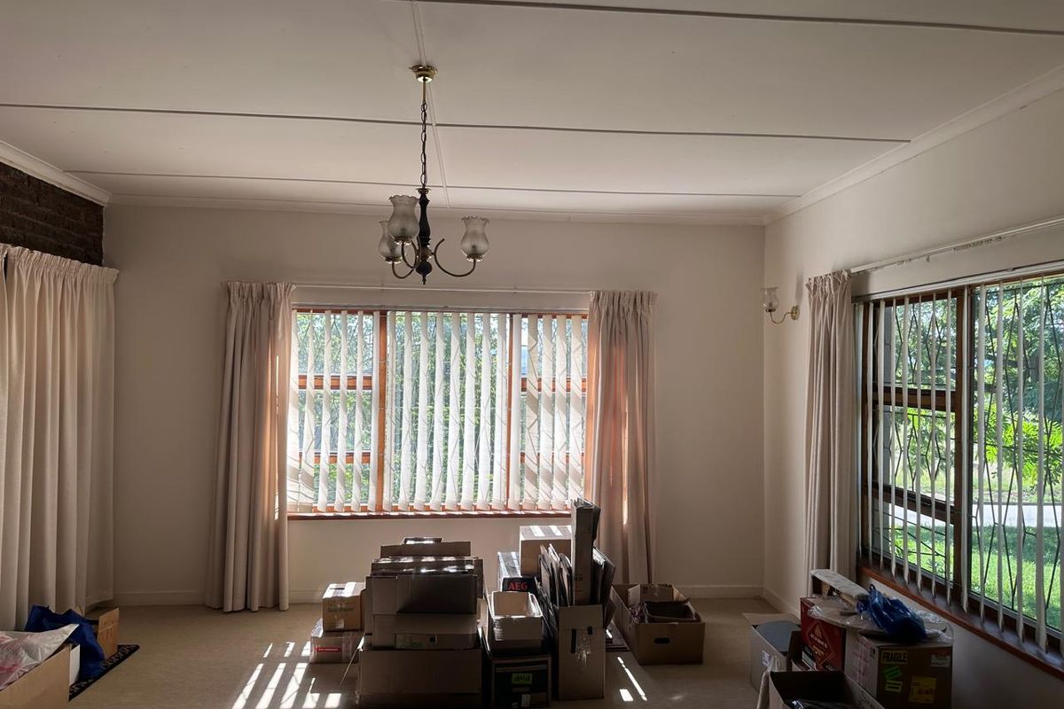 A spacious, light-filled living room featuring large windows with vertical blinds, security bars, and a classic ceiling chandelier.