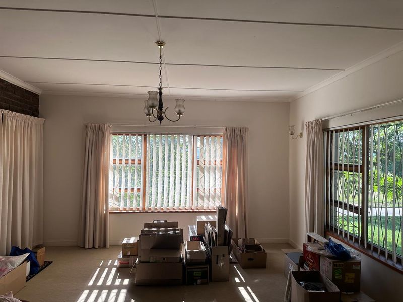 A spacious, light-filled living room featuring large windows with vertical blinds, security bars, and a classic ceiling chandelier.