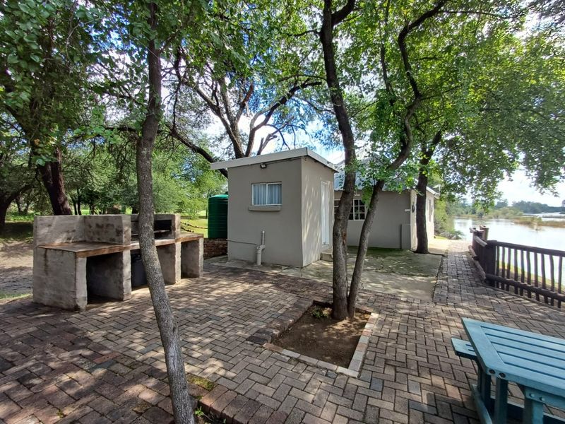 Outdoor entertainment area featuring a built-in braai, brick paving, and a water tank, set in a scenic riverside garden environment.