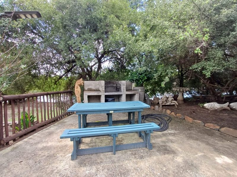 Outdoor patio area featuring a built-in masonry braai, concrete flooring, and timber railing, set within a natural garden environment.
