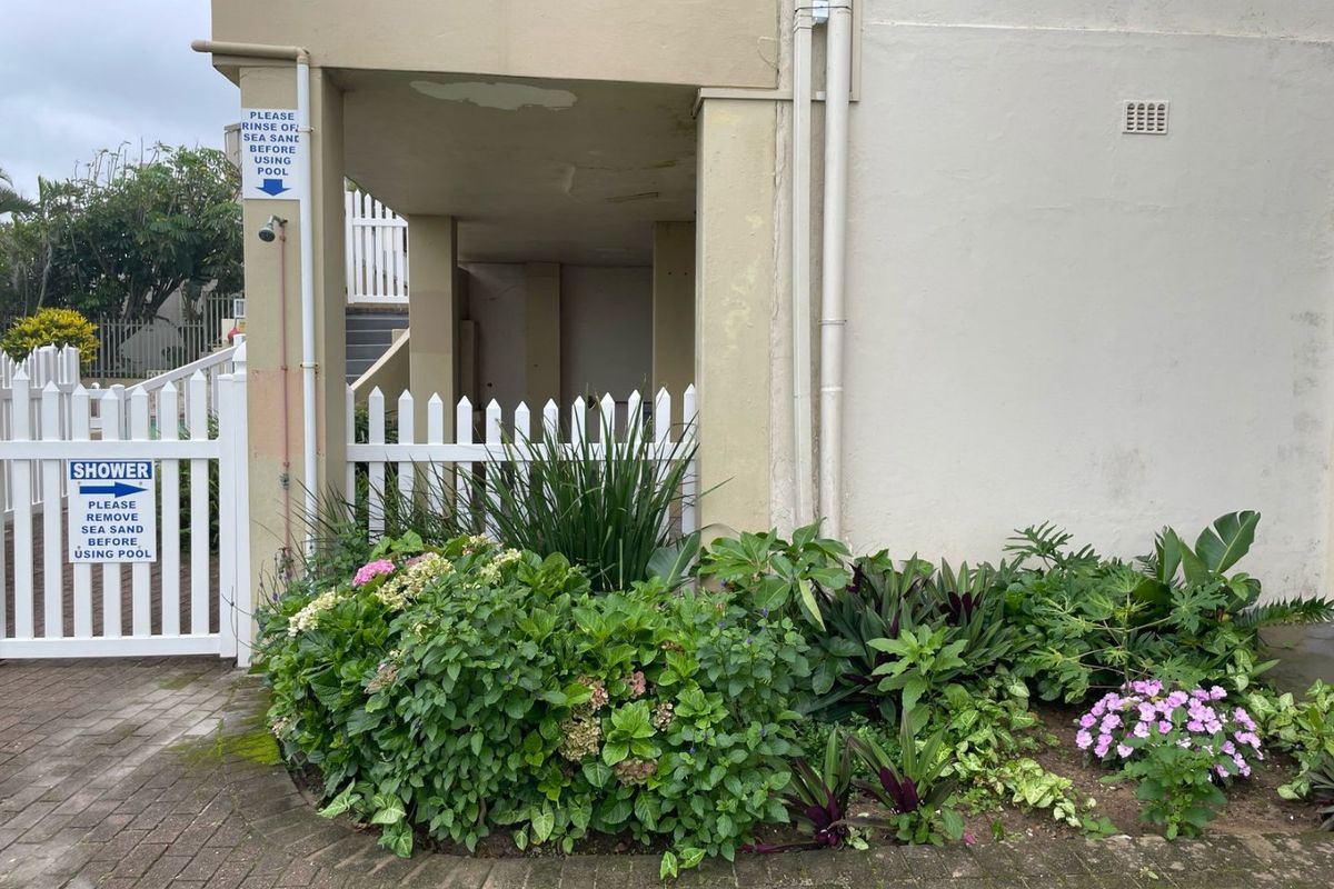 Exterior view of a building corner featuring a landscaped garden, paved walkway, white picket fence, and a dedicated outdoor shower area.