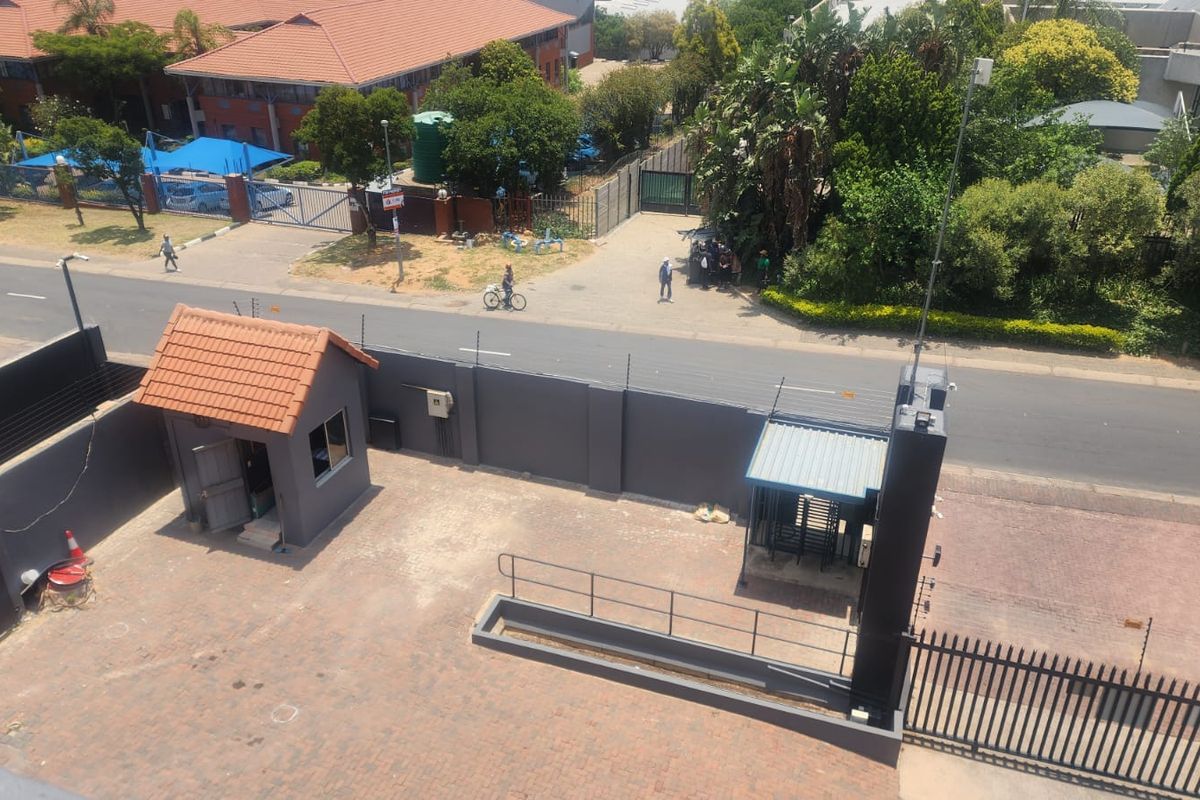 Aerial view of a commercial property entrance featuring a security guard house, perimeter wall with electric fencing, and paved access area.